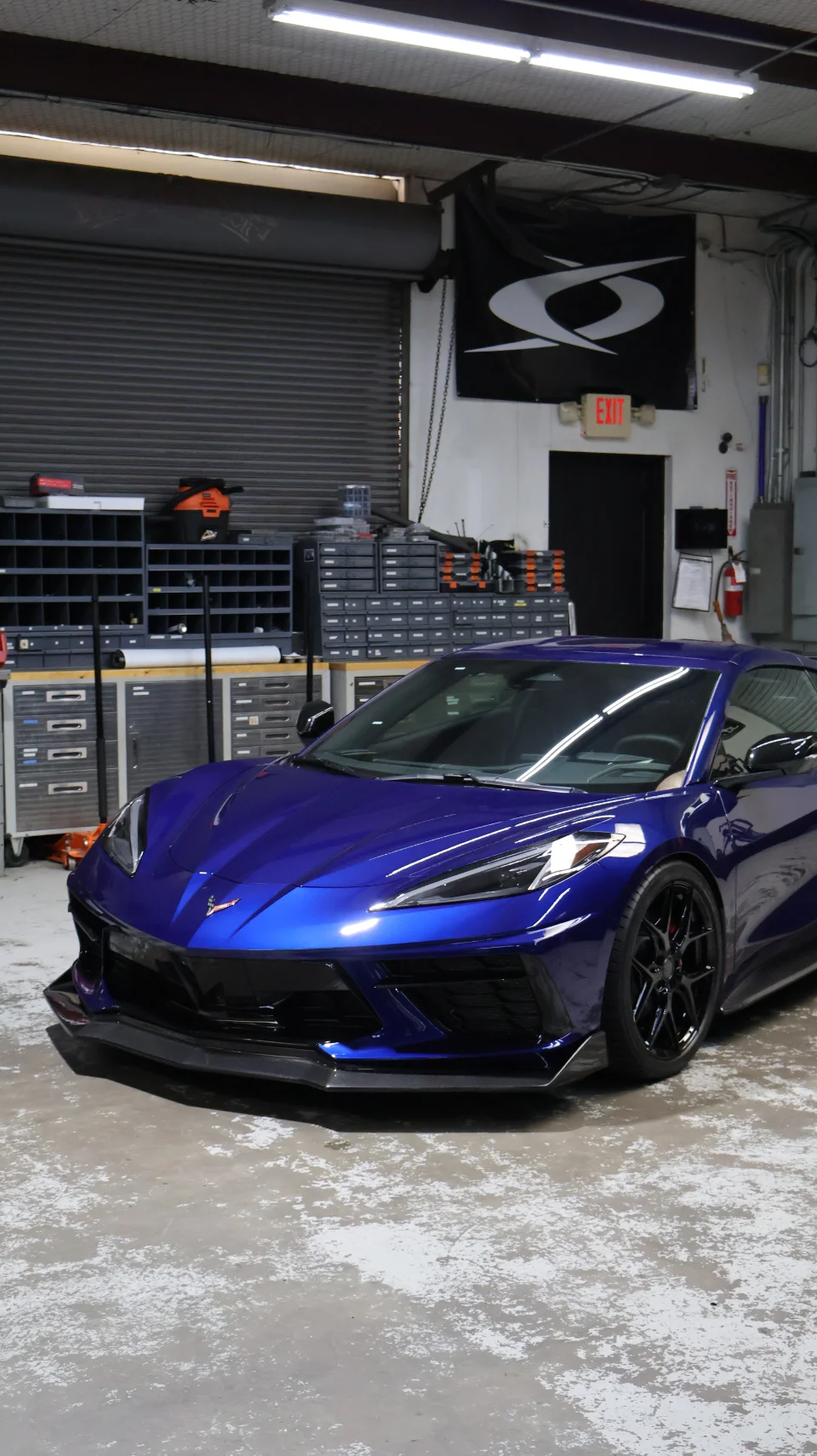 A vibrant blue Chevrolet Corvette C8 with a front splitter parked inside a well-lit performance shop.