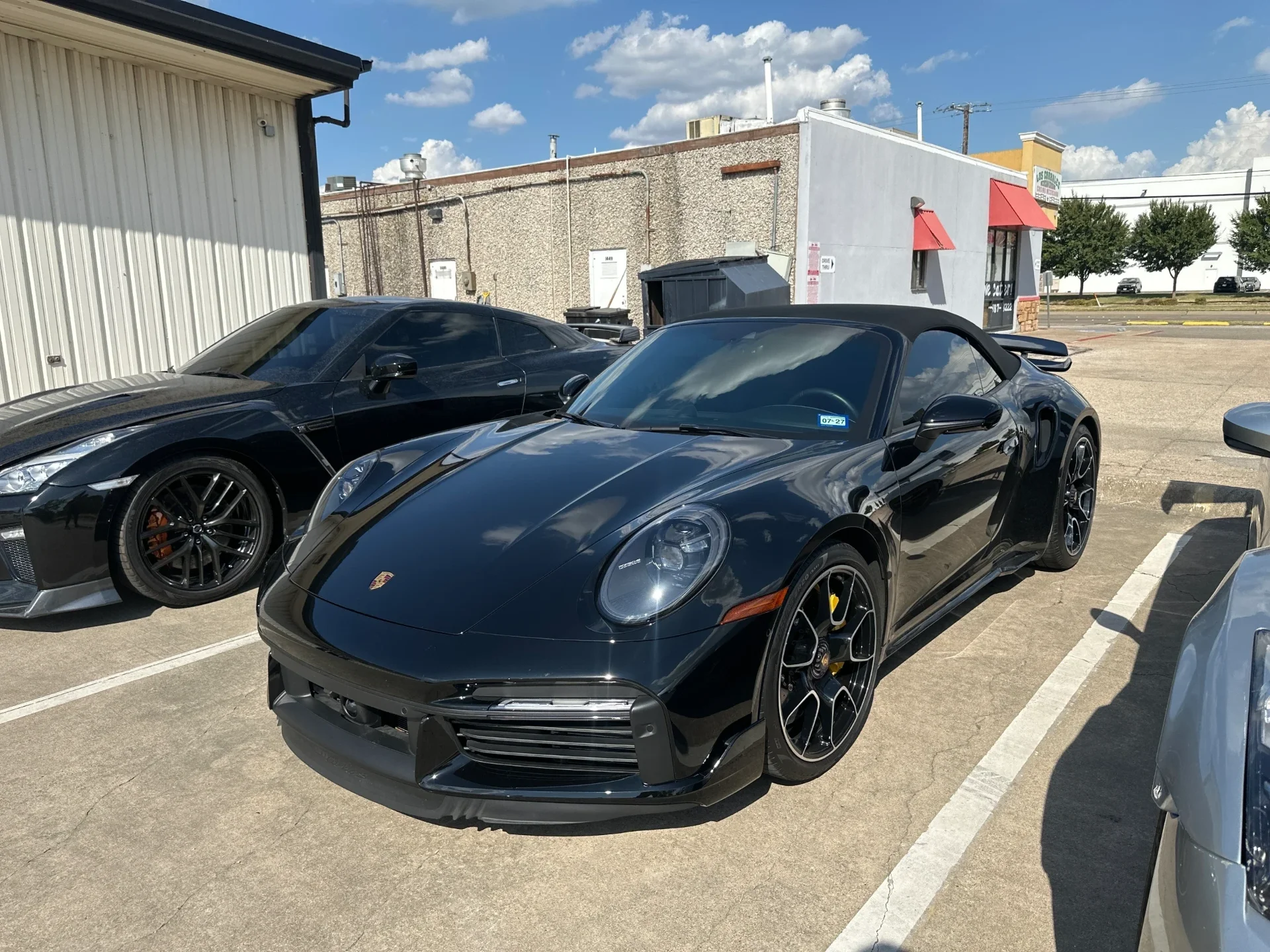 A black Porsche 911 992 Turbo S Cabriolet parked next to a black Nissan GT-R R35 in a sunny parking lot.