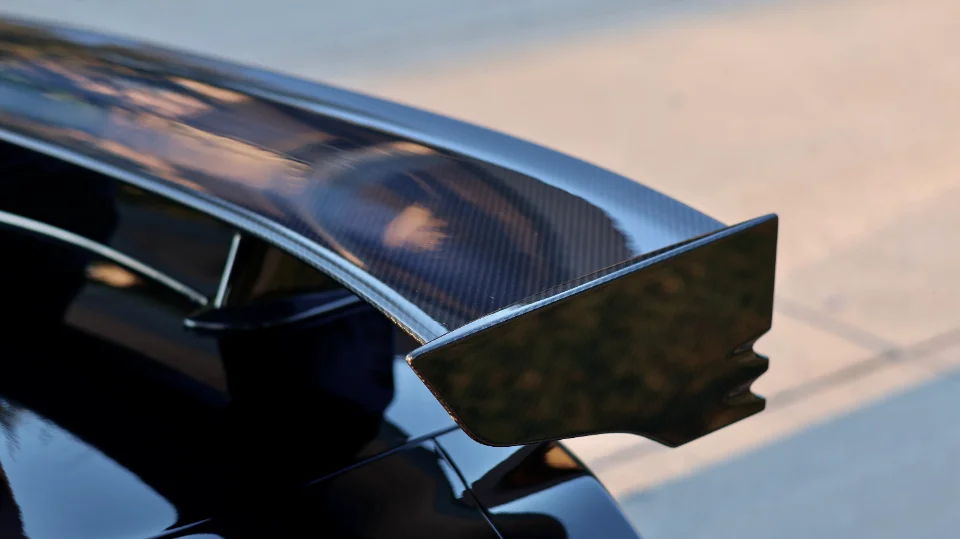 Close-up of a glossy carbon fiber rear wing on a performance car, showcasing its weave and aerodynamic shape.