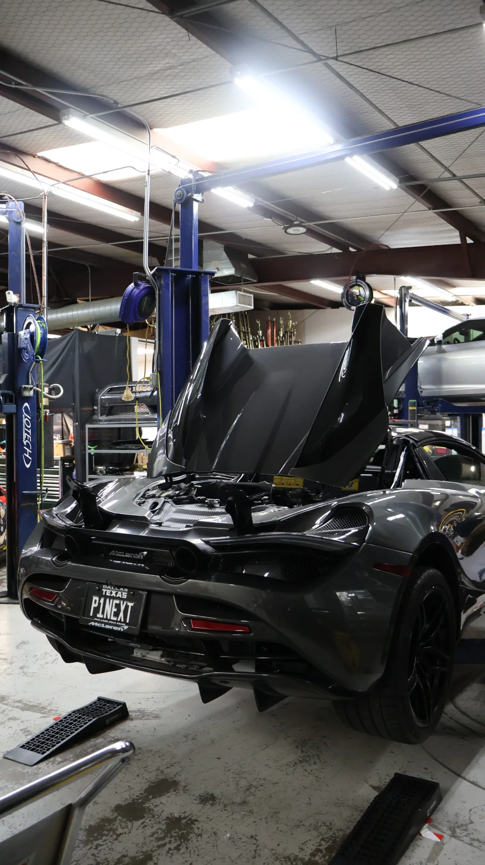A grey McLaren 720S with its engine bay open, on a car lift in a performance tuning shop.