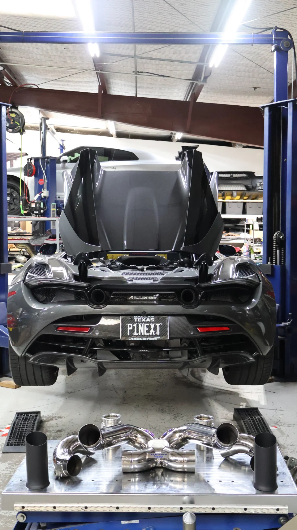 A grey McLaren 720S on a lift in a performance shop with its engine bay open, an aftermarket titanium exhaust system displayed on a table.