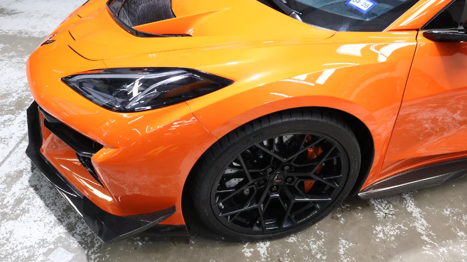 Close-up of an orange Corvette C8 Z06 featuring a carbon fiber front splitter, black multi-spoke wheels, and orange brake calipers.