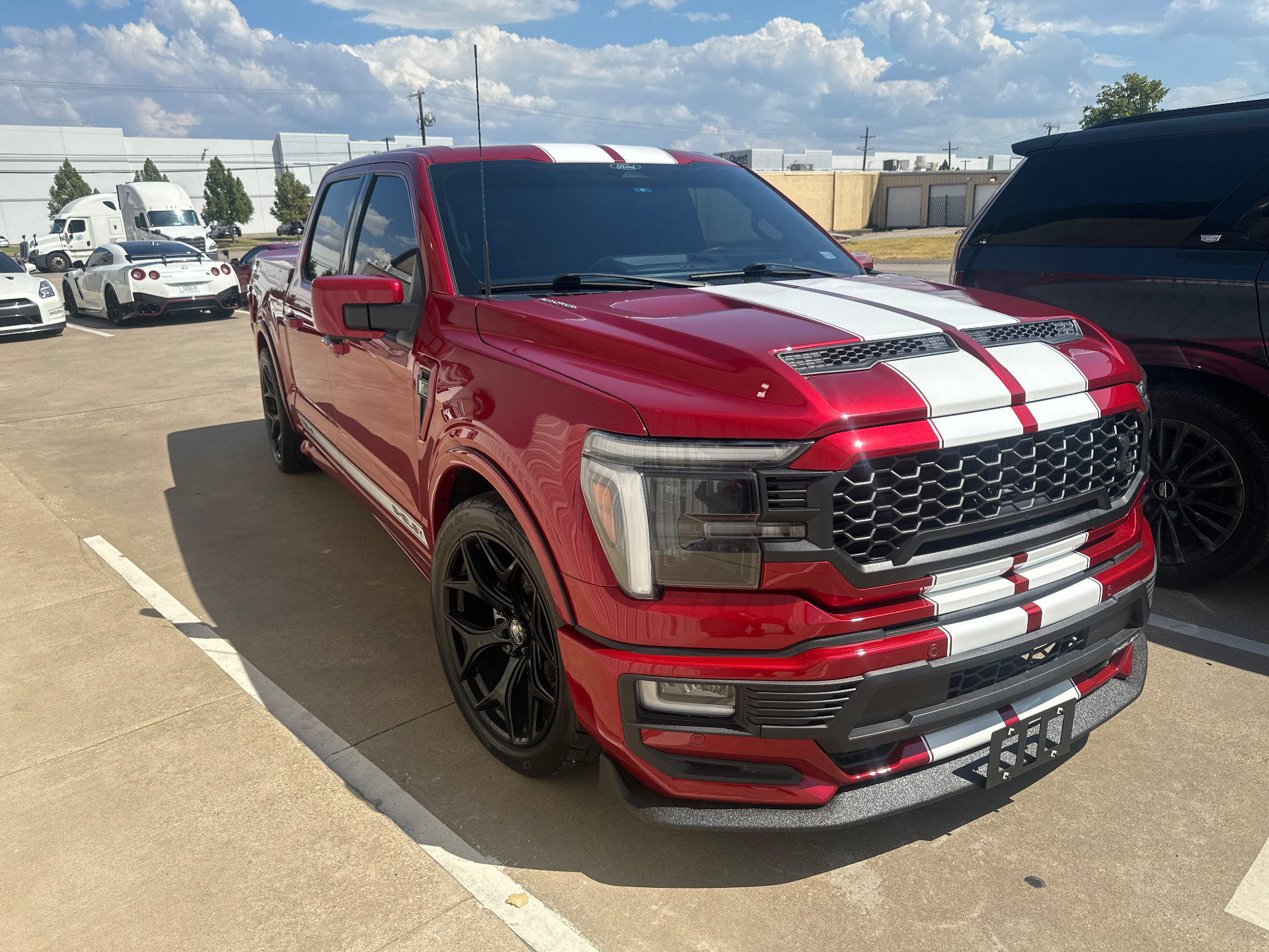 A red Ford F-150 Shelby truck with white racing stripes parked in a lot with a white Nissan GT-R in the background.