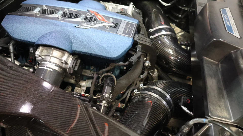 A close-up of a C8 Corvette engine bay featuring a blue engine cover and carbon fiber aftermarket induction system.