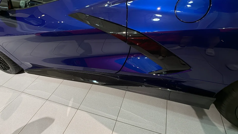Close-up of the side of a blue Corvette C8, showcasing a carbon fiber side skirt in a showroom setting.