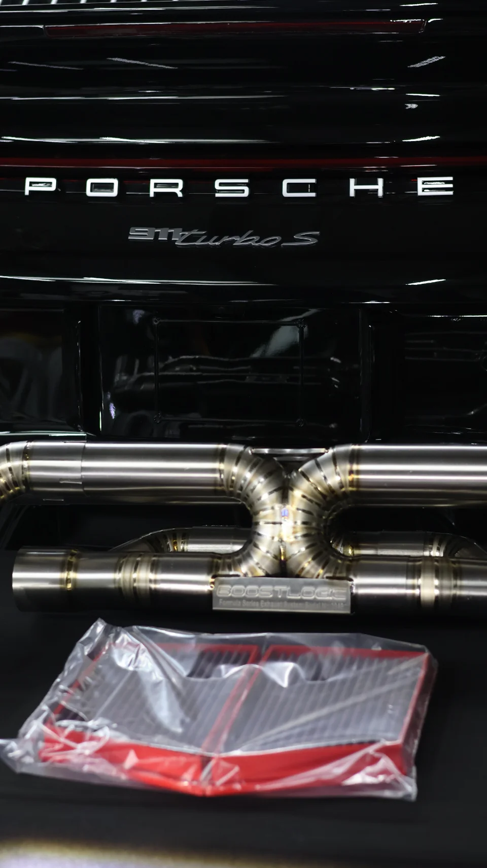 Close-up of a Boost Logic titanium exhaust system installed on a black Porsche 911 Turbo S, with an aftermarket air filter in packaging.