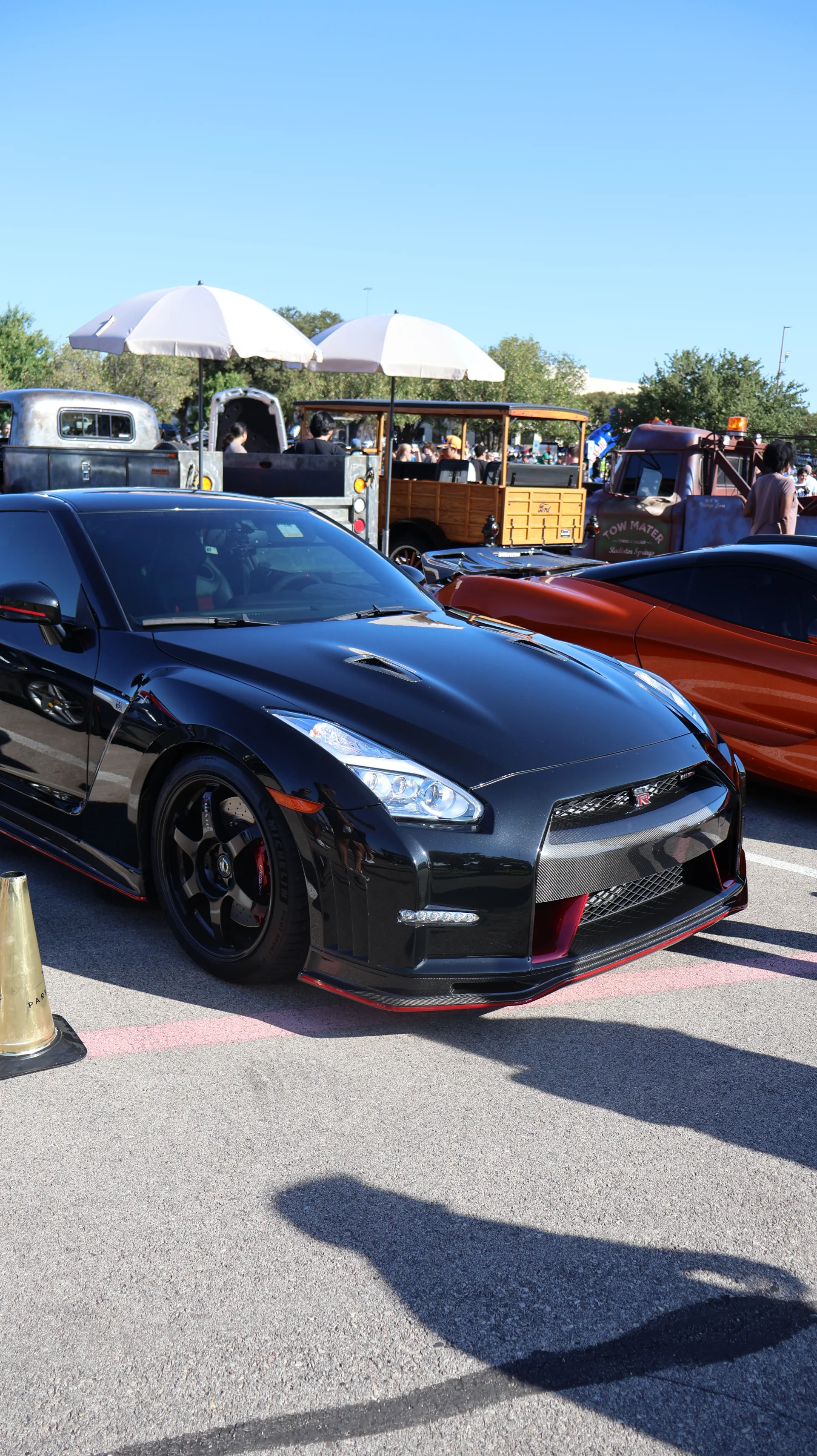 A black Nissan GT-R with red accents and aftermarket wheels parked at an outdoor car show under a clear blue sky.