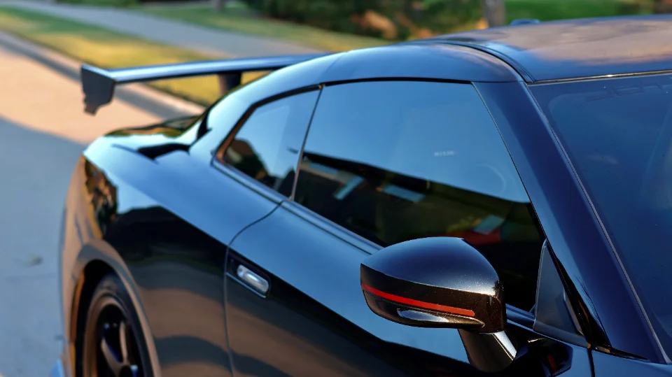 Close-up of a black Nissan GT-R's rear quarter panel, featuring a large aftermarket wing and side mirror with red accent.