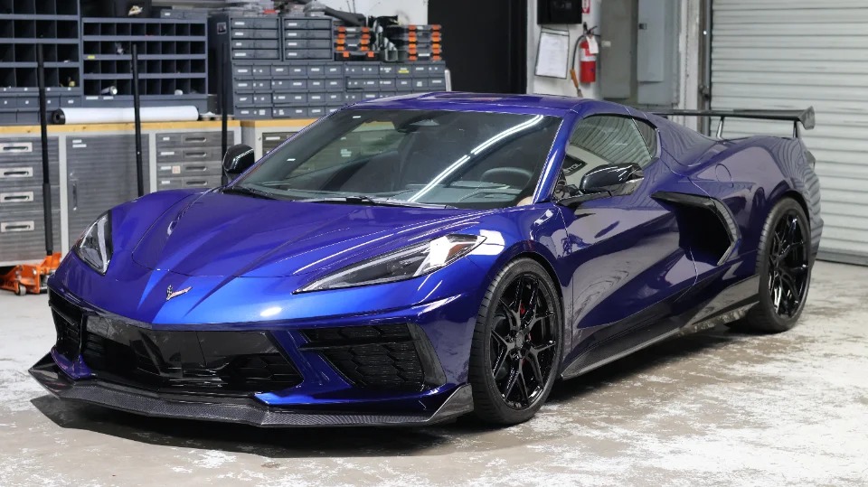 A blue Chevrolet Corvette C8 with carbon fiber aerodynamic parts and aftermarket wheels parked inside a performance shop.