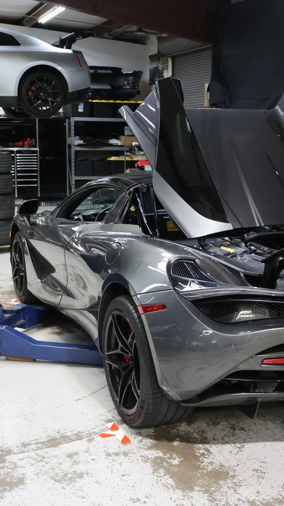 A grey McLaren 720S with its hood and door open, on a lift in a performance tuning shop, with a Nissan GT-R in the background.