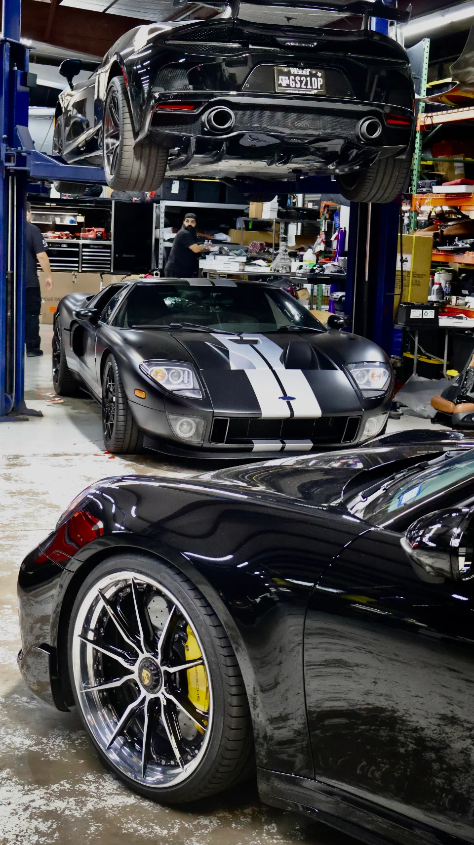 Three high-performance cars, a McLaren, Ford GT, and Porsche, inside a performance tuning shop with one car on a lift.