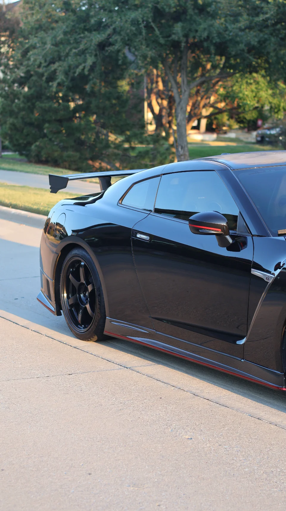 A black Nissan GT-R with aftermarket wheels, carbon fiber side skirts, and a large rear wing parked outdoors.