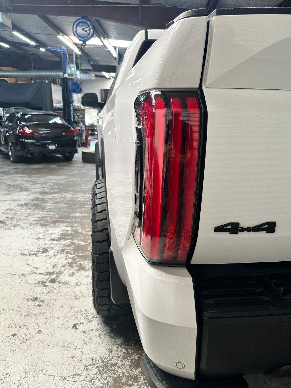Rear view of a white pickup truck with aftermarket LED taillights and large off-road tires, parked inside a car shop.