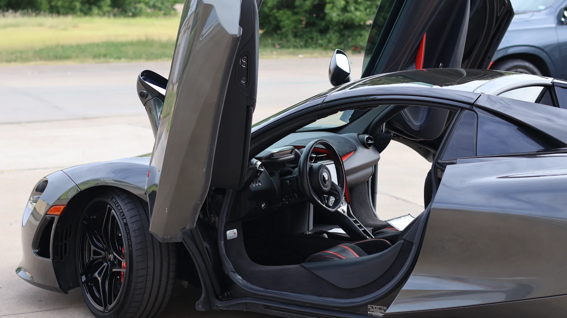 A grey McLaren 720S supercar with its butterfly doors open, showcasing the black and red interior.