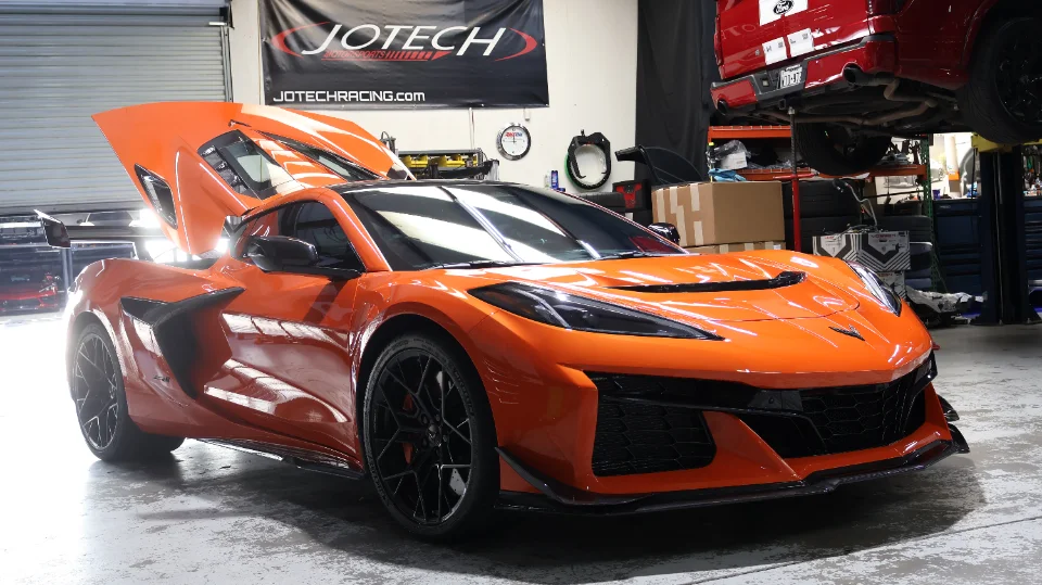 An orange Chevrolet Corvette C8 Z06 with its engine cover open, parked inside the Jotech Racing performance shop.