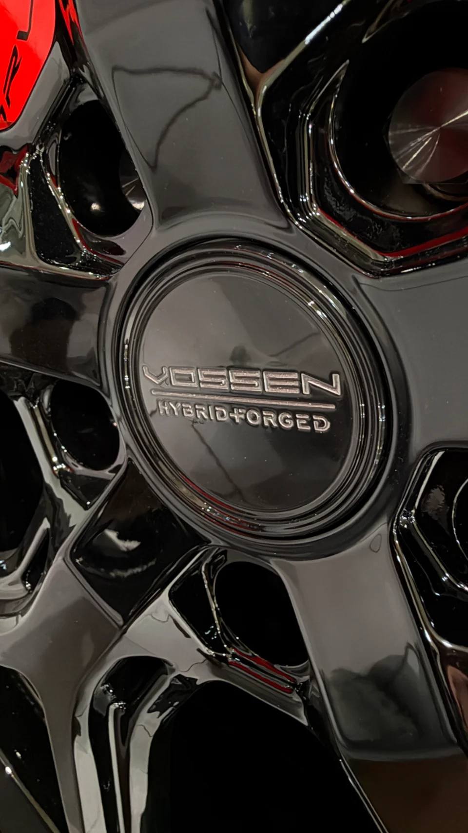 Close-up detail of a glossy black Vossen Hybrid Forged wheel with a red brake caliper visible in the background.