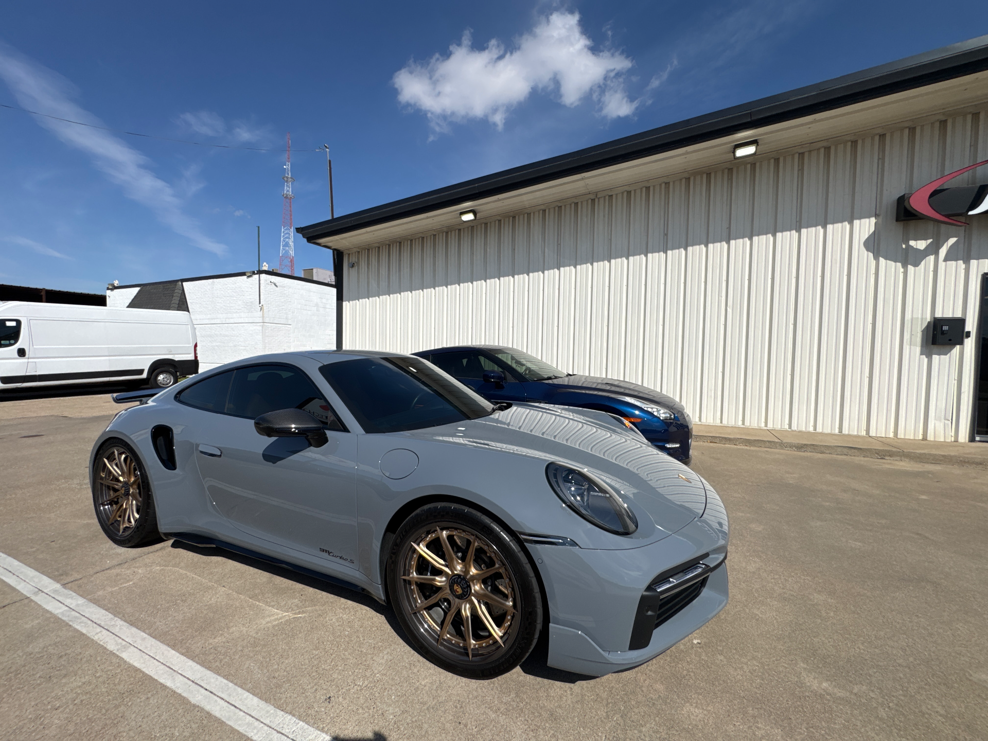 A sleek grey Porsche 911 with bronze wheels parked outside a performance shop, with a blue sports car in the background.