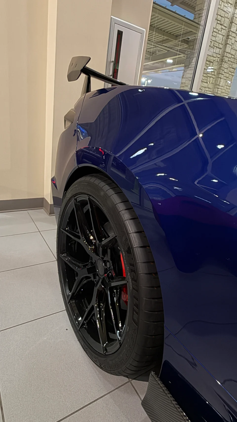 Close-up of a blue car's rear wheel featuring black multi-spoke rims, red brake calipers, and a carbon fiber aero element.