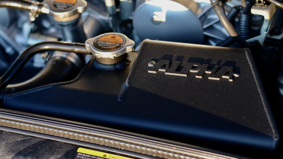 Close-up of an Alpha Performance intake manifold and radiator cap in a car's engine bay, showcasing performance parts.