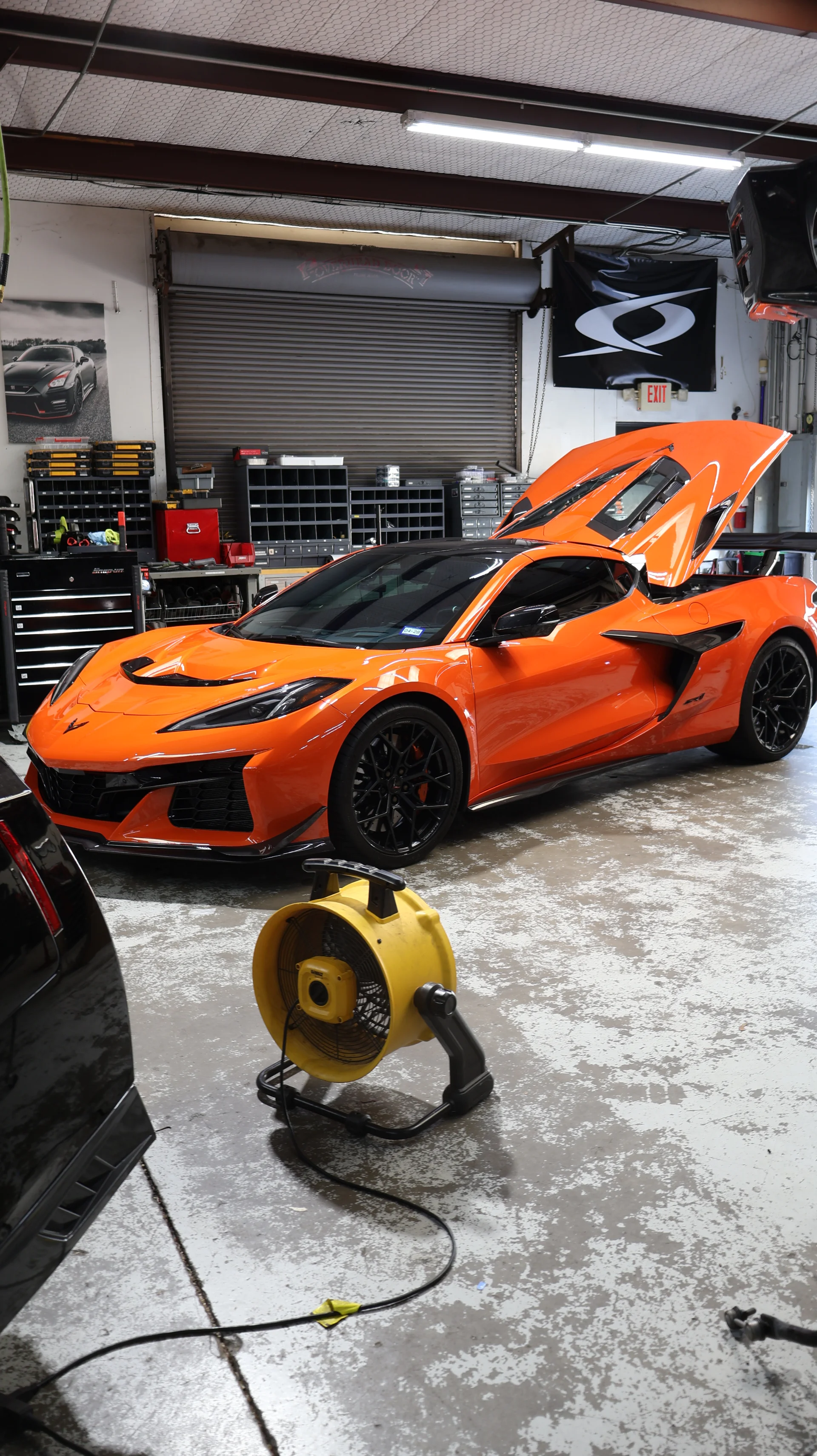 An orange Corvette Z06 with its hood and engine cover open, parked inside a performance tuning shop.