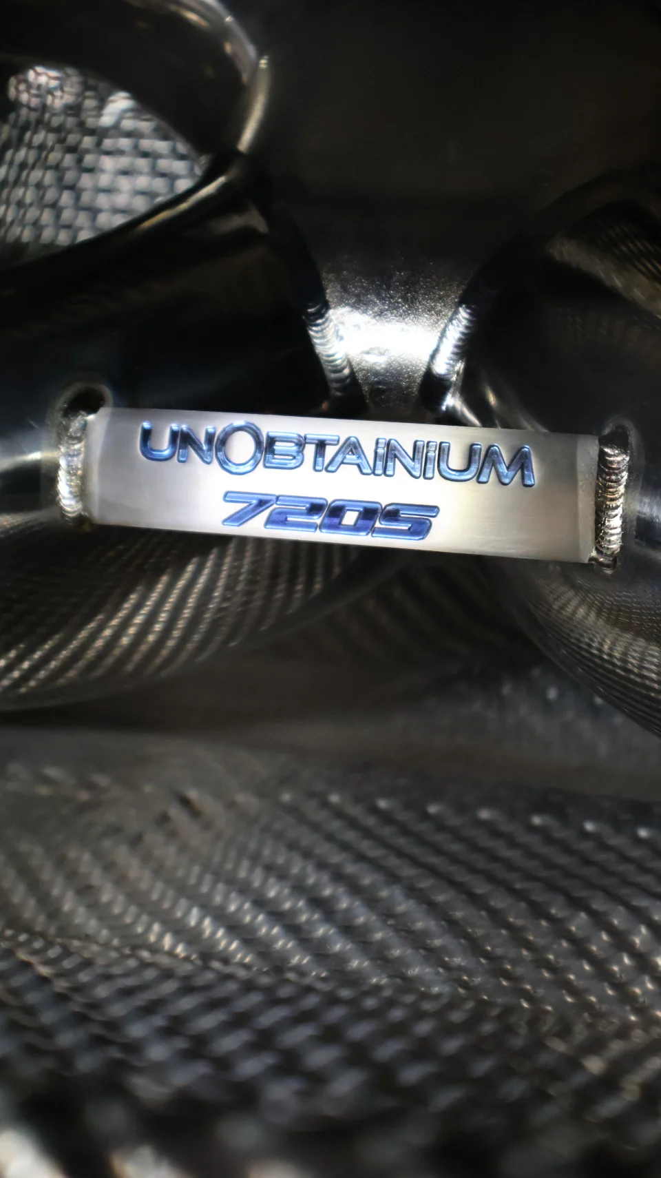 Close-up detail of an Unobtainium exhaust component for a McLaren 720S, showing the engraved logo and welds.