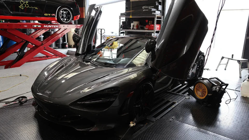 A grey McLaren 720S with butterfly doors open, strapped to a dynamometer in a performance tuning shop.