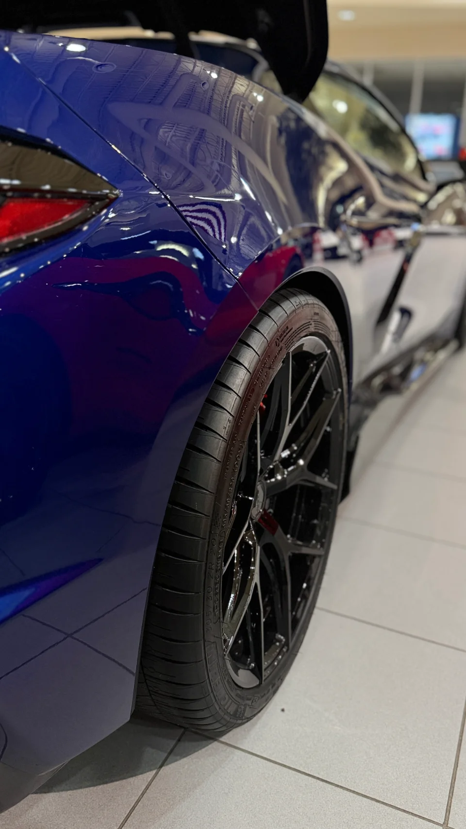 Close-up detail of the rear wheel and fender of a blue Chevrolet Corvette C8 Z06, showcasing the Michelin tire and black multi-spoke wheel in a showroom.