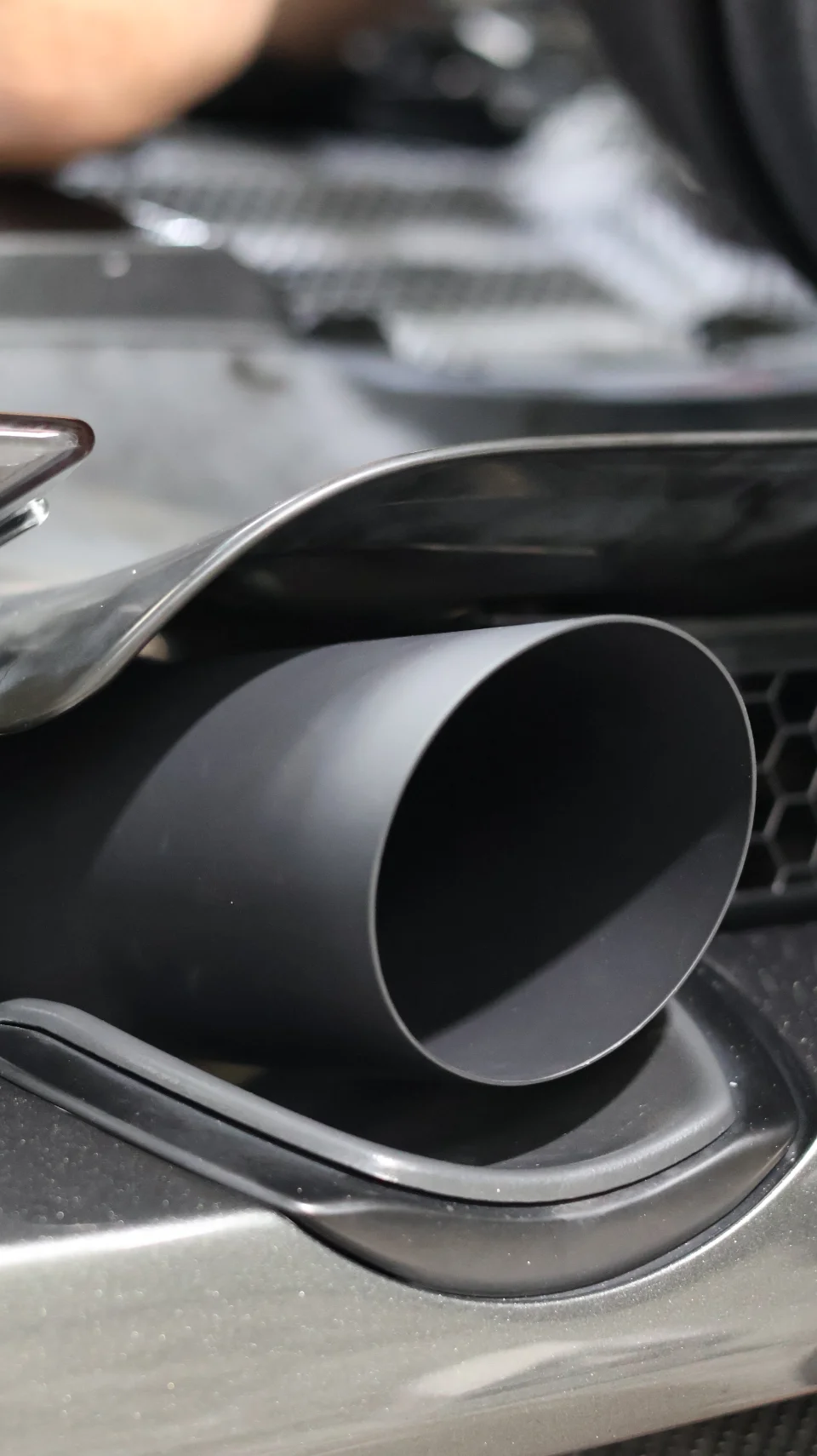 A close-up detail shot of a matte black exhaust tip integrated into a car's rear bumper with a honeycomb grille.