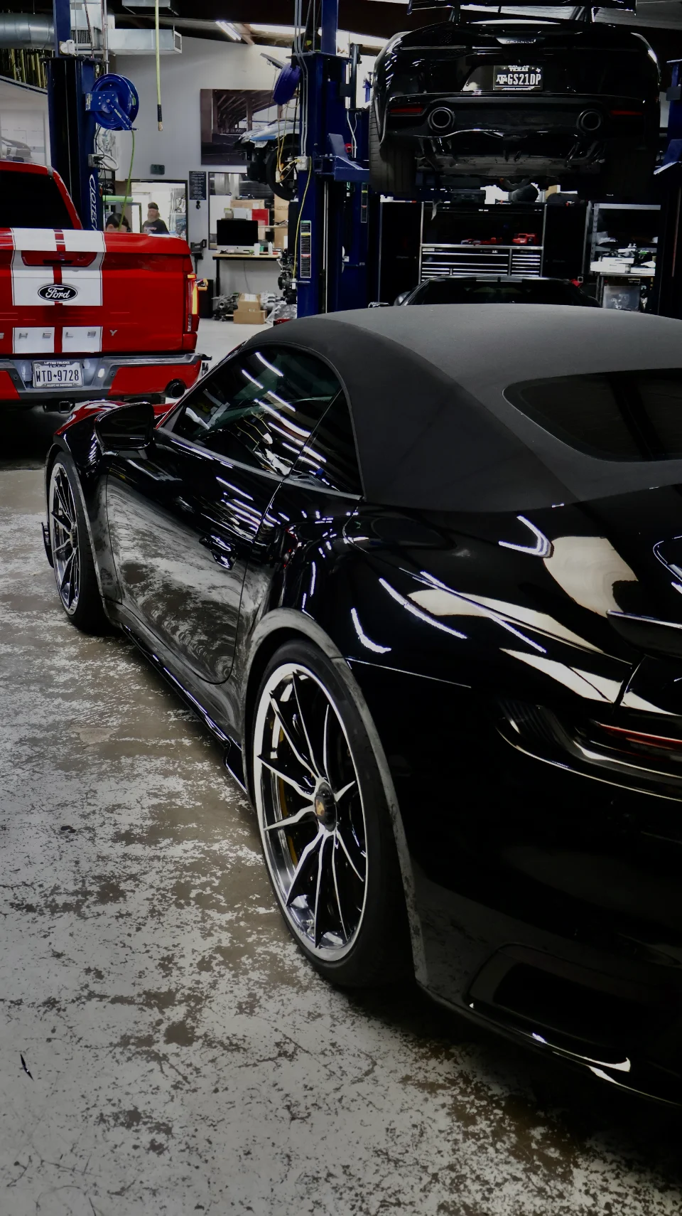 A black Porsche 911 convertible parked in a performance tuning shop with other vehicles on lifts in the background.