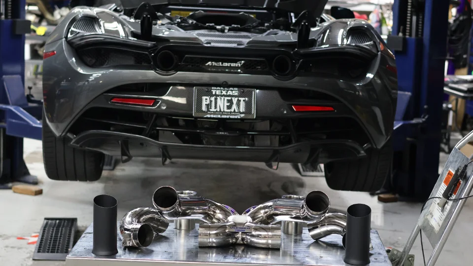 A grey McLaren sports car on a lift in a shop, with a new polished performance exhaust system displayed on a table below.