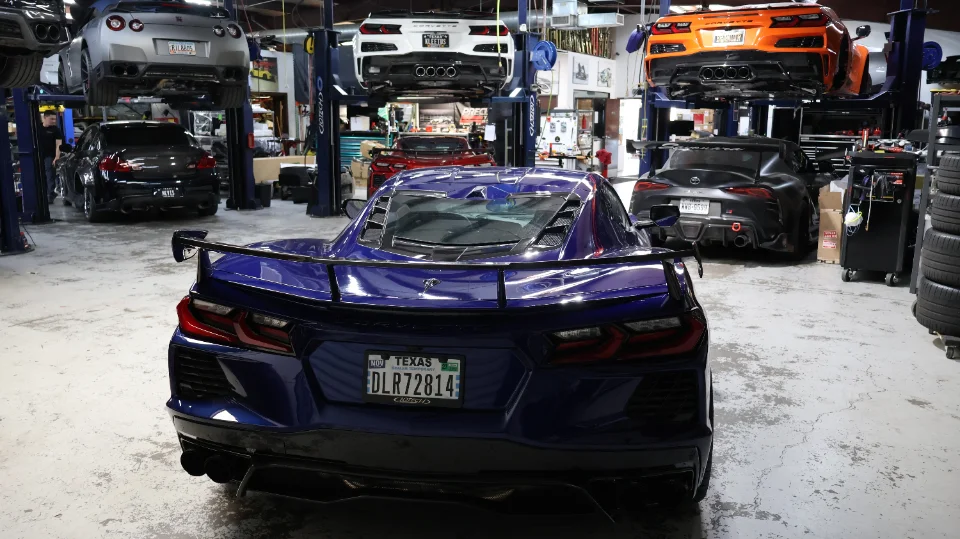 A blue Corvette C8 in the foreground of a busy performance tuning shop with multiple other sports cars on lifts.