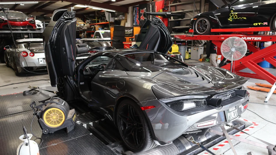 A grey McLaren 720S with its doors open, strapped to a dynamometer in a performance tuning shop.