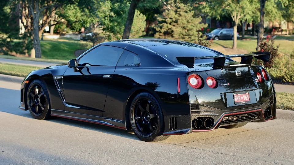 A black Nissan GT-R with aftermarket wheels and a carbon fiber wing parked on a sunny street.