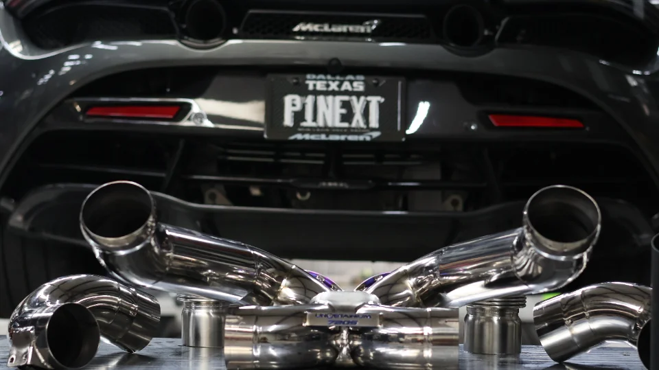 Aftermarket titanium exhaust system laid out in front of a McLaren supercar with a custom P1NEXT license plate.