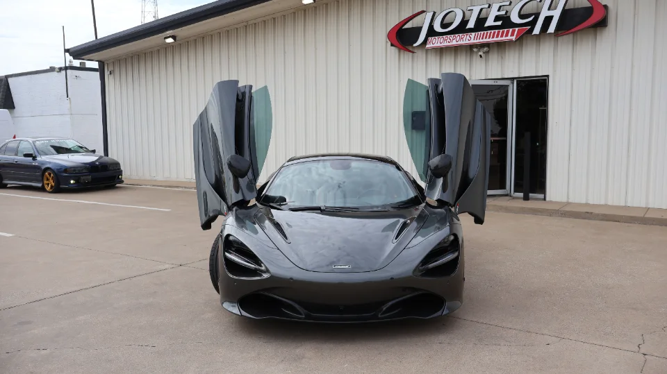 A grey McLaren 720S with its butterfly doors open, parked prominently in front of the Jotech Motorsports shop.