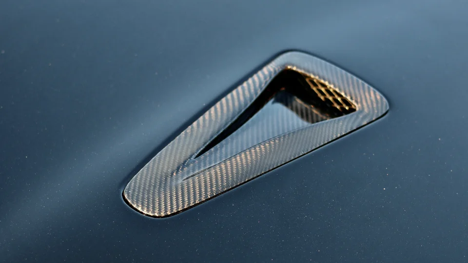 Close-up of a carbon fiber hood vent on a dark car's hood, showcasing the weave pattern and aerodynamic design.