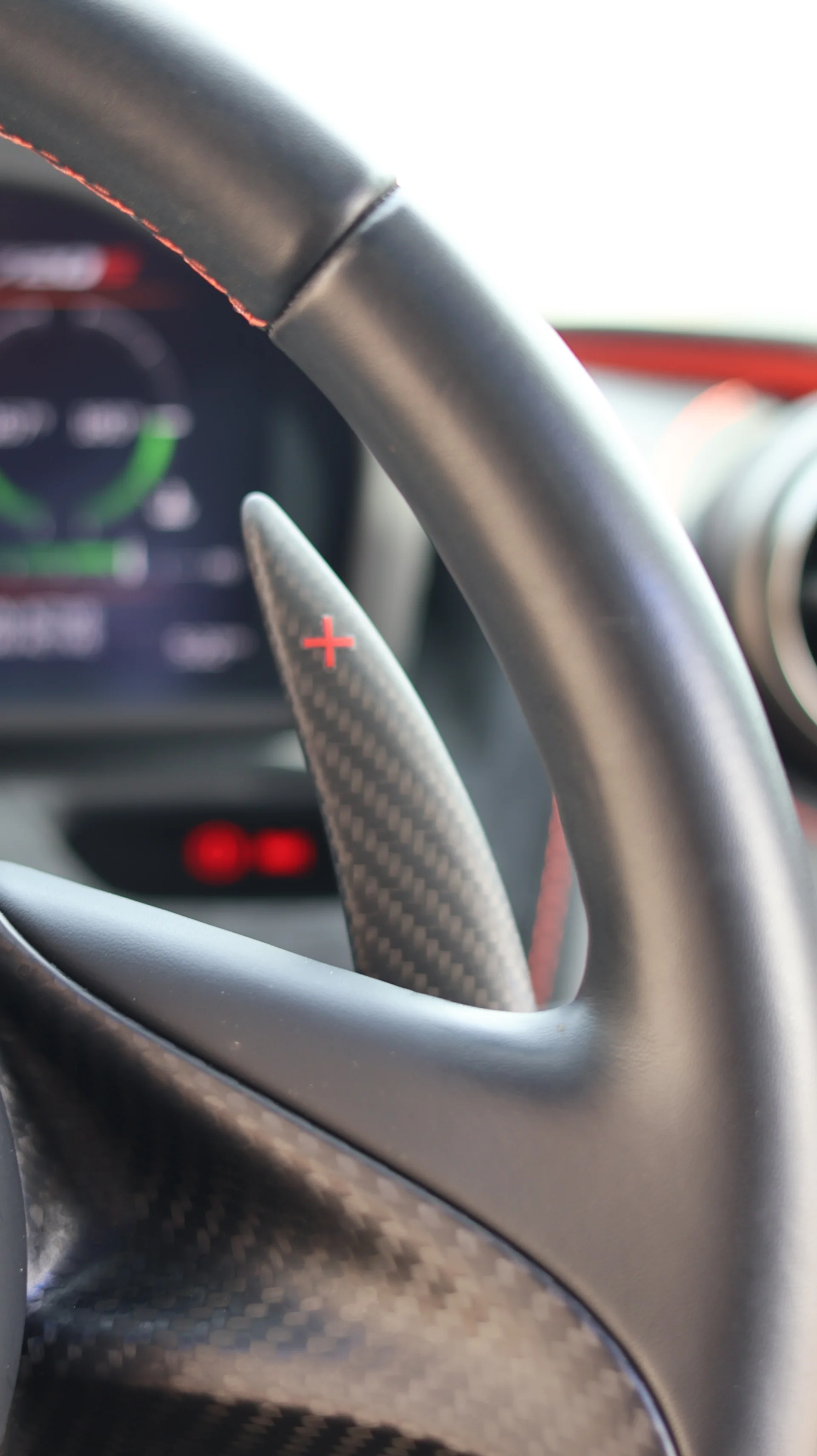 Close-up of a carbon fiber paddle shifter with a red plus sign on a steering wheel, with a digital dashboard in the background.