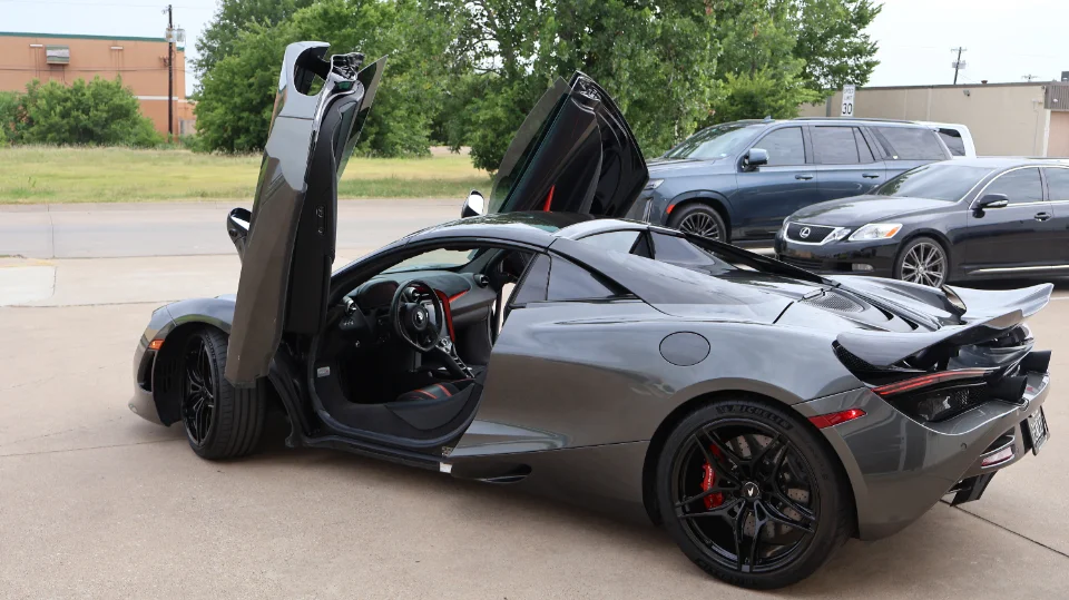 A grey McLaren 720S supercar parked outdoors with its butterfly doors open, showcasing its sleek design and interior.