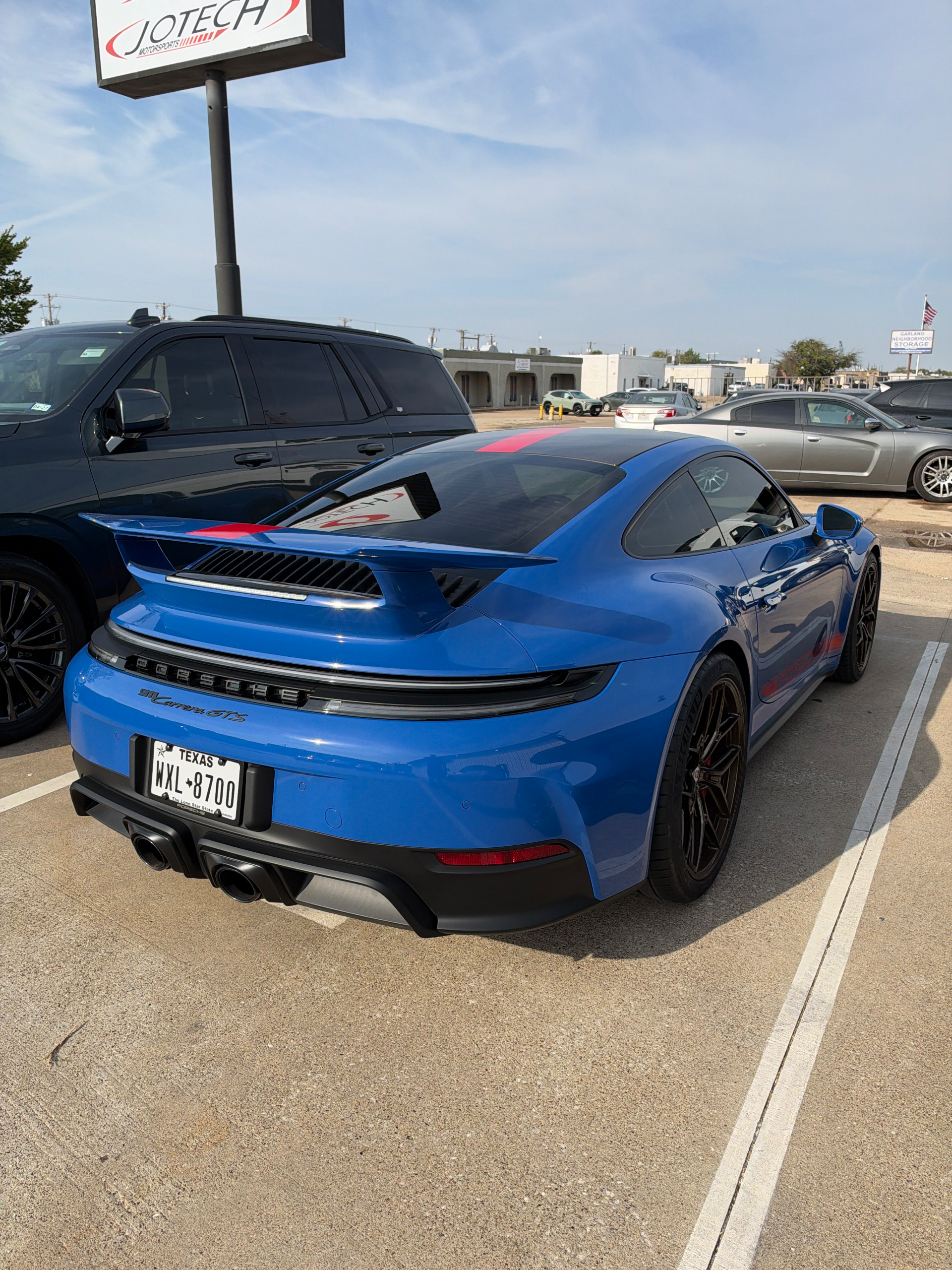 Rear view of a blue Porsche 911 Carrera GTS with red racing stripes parked in a Jotech Motorsports parking lot.
