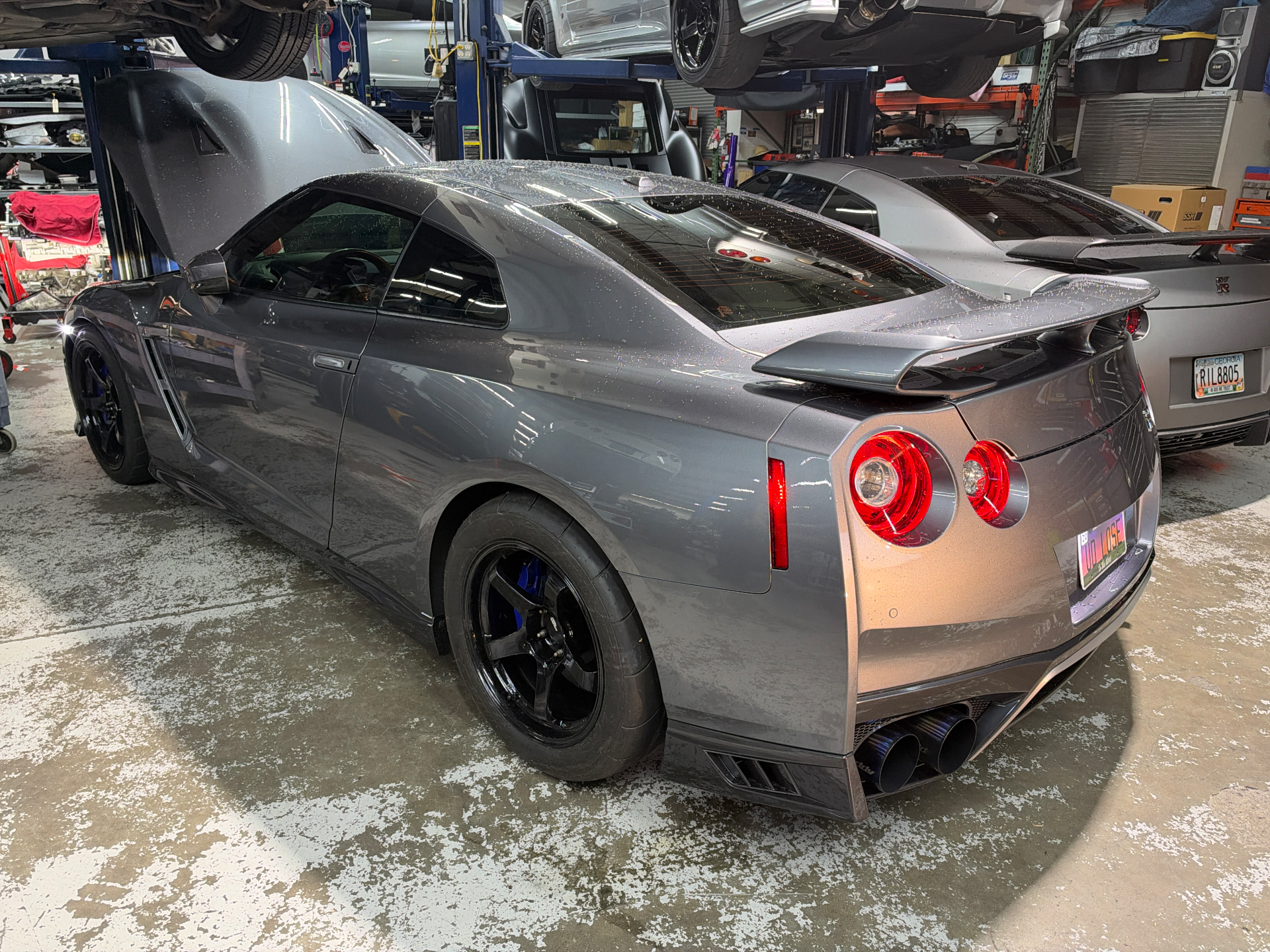 A grey Nissan GT-R with its hood open, parked in a performance tuning shop with other cars on lifts.
