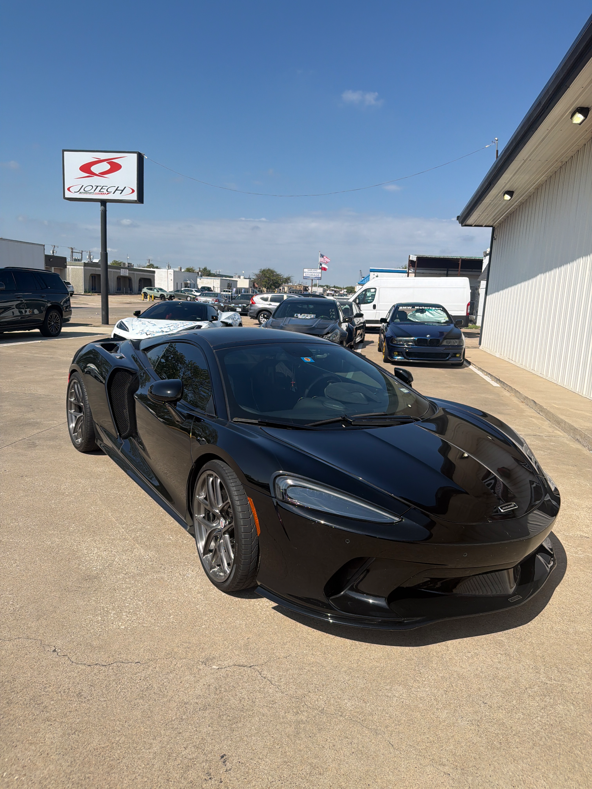 A sleek black McLaren supercar is parked outside the Jotech Motorsports facility under a clear blue sky.