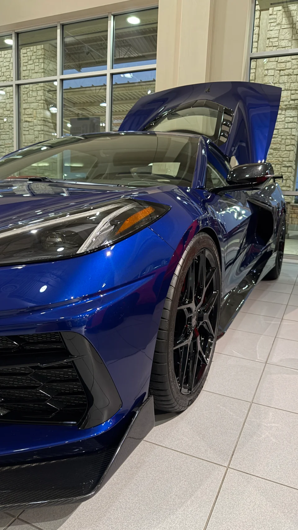 A blue C8 Corvette with its engine cover open, showcasing carbon fiber aerodynamic elements in a showroom.
