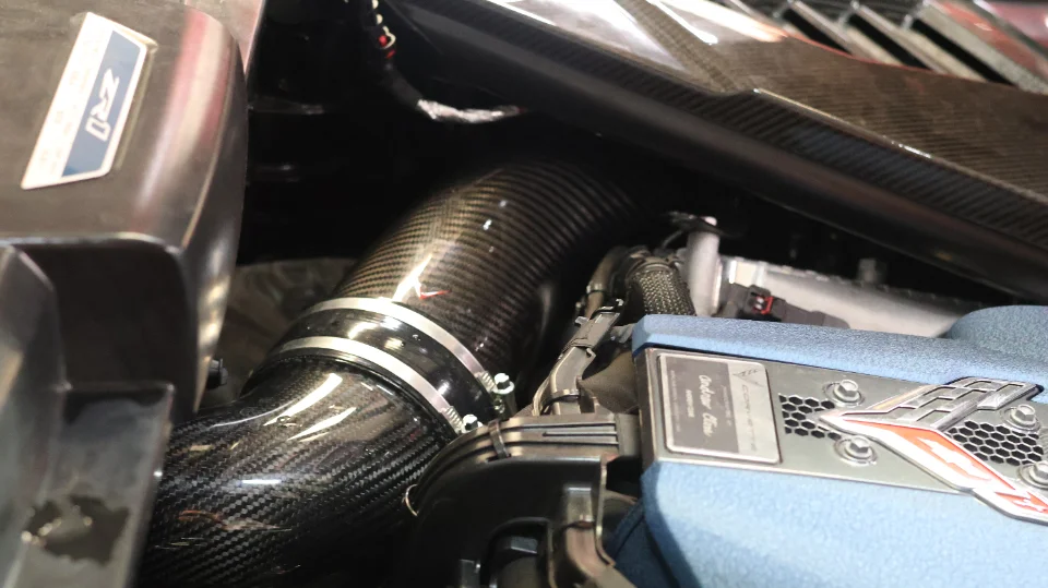 A close-up of a C8 Corvette engine bay featuring a carbon fiber induction system and blue engine cover.