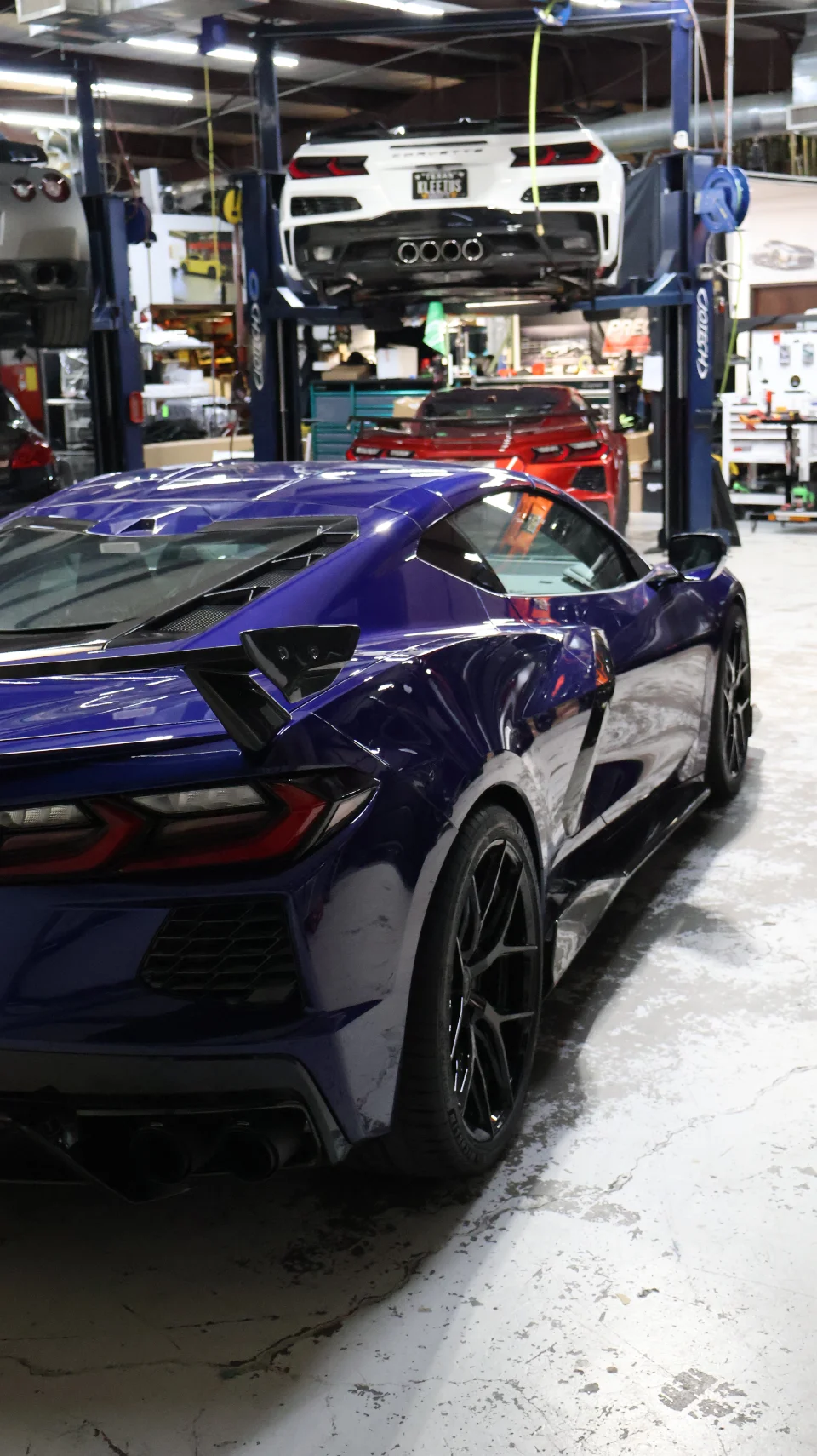 A blue Corvette C8 parked in a performance tuning shop with a white Corvette C8 on a lift in the background.