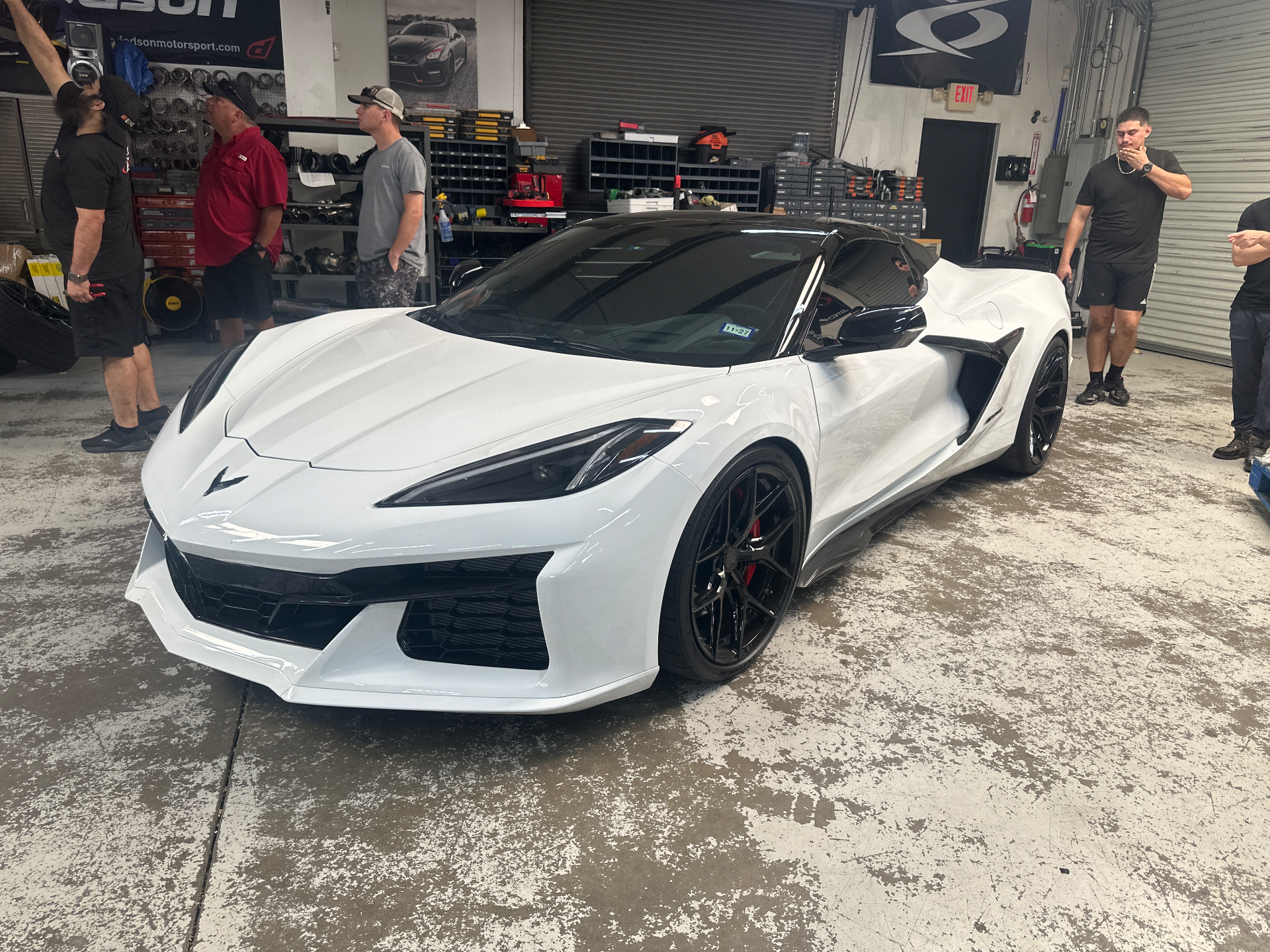 A white Chevrolet Corvette C8 with black aftermarket wheels parked inside a performance tuning shop, with several people standing nearby.