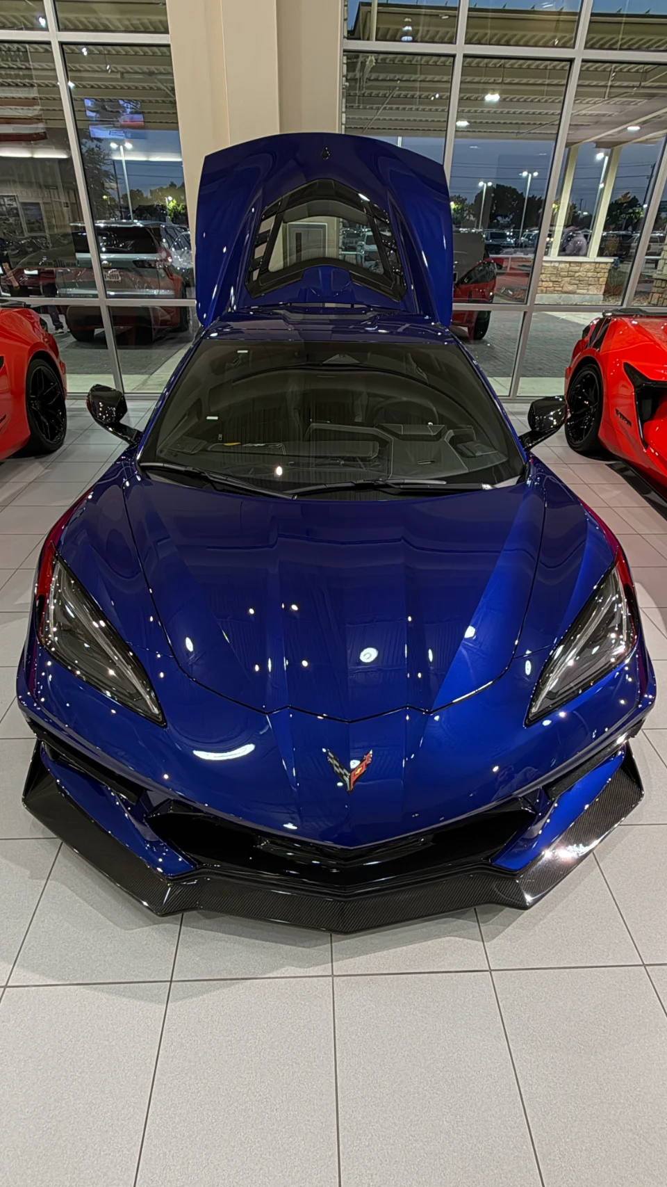 A blue Chevrolet Corvette C8 with its engine bay open, displayed in a car dealership showroom.