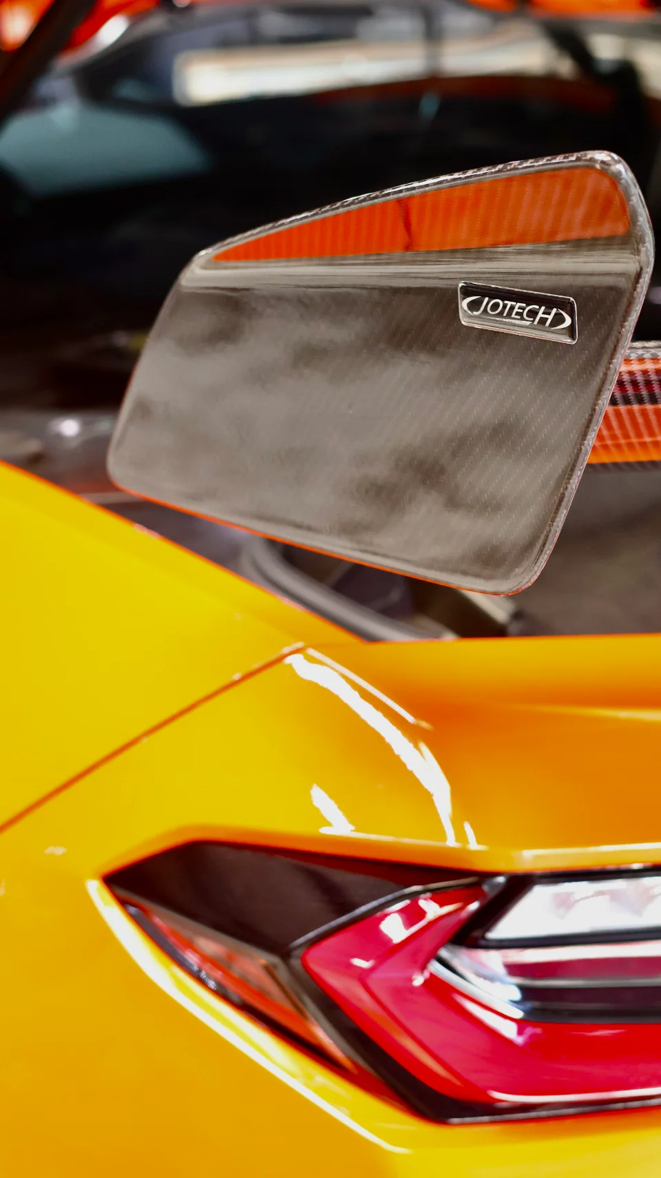 Close-up of an orange car's carbon fiber exterior component featuring a Jotech logo and a red light detail.