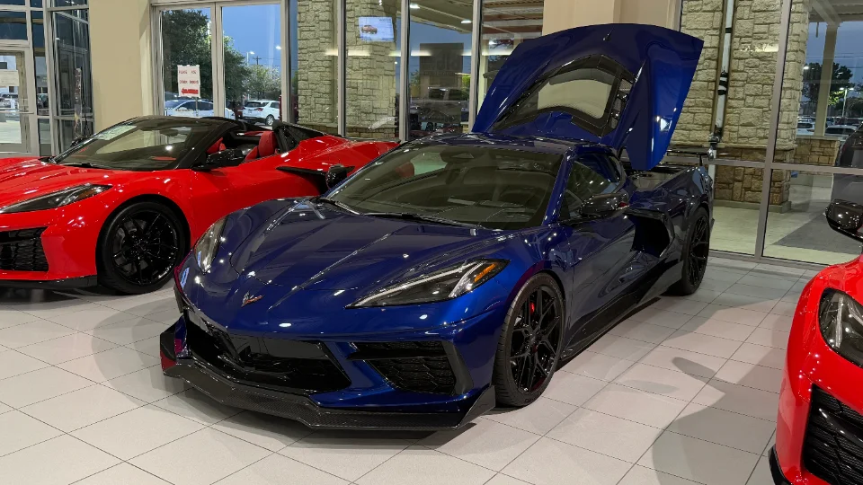 A blue Chevrolet Corvette C8 Z06 with its engine bay open, parked next to a red Corvette in a dealership showroom.