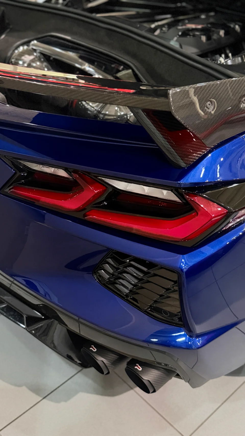 A close-up rear view of a blue Chevrolet Corvette C8, highlighting its carbon fiber spoiler, LED taillights, and performance exhaust tips.