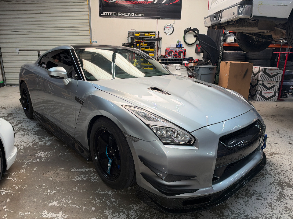 A silver Nissan GT-R with carbon fiber aero and aftermarket wheels parked inside a performance tuning shop.
